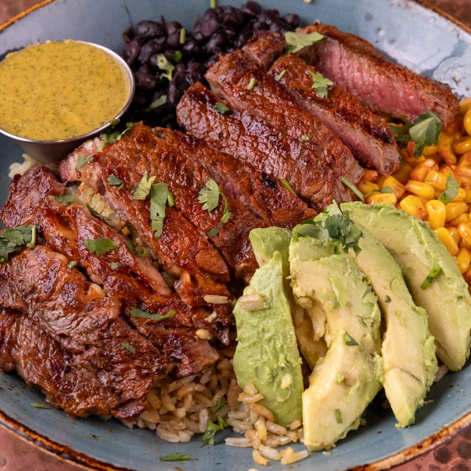 A Mexican steak bowl with rice, black beans, corn, avocado slices, and a side of green sauce.