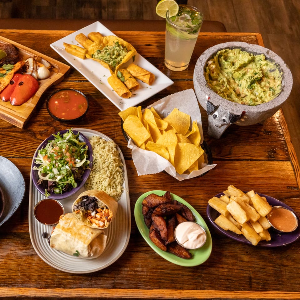 A wooden table laden with various Mexican dishes, including burritos, guacamole, chips, and a cocktail.