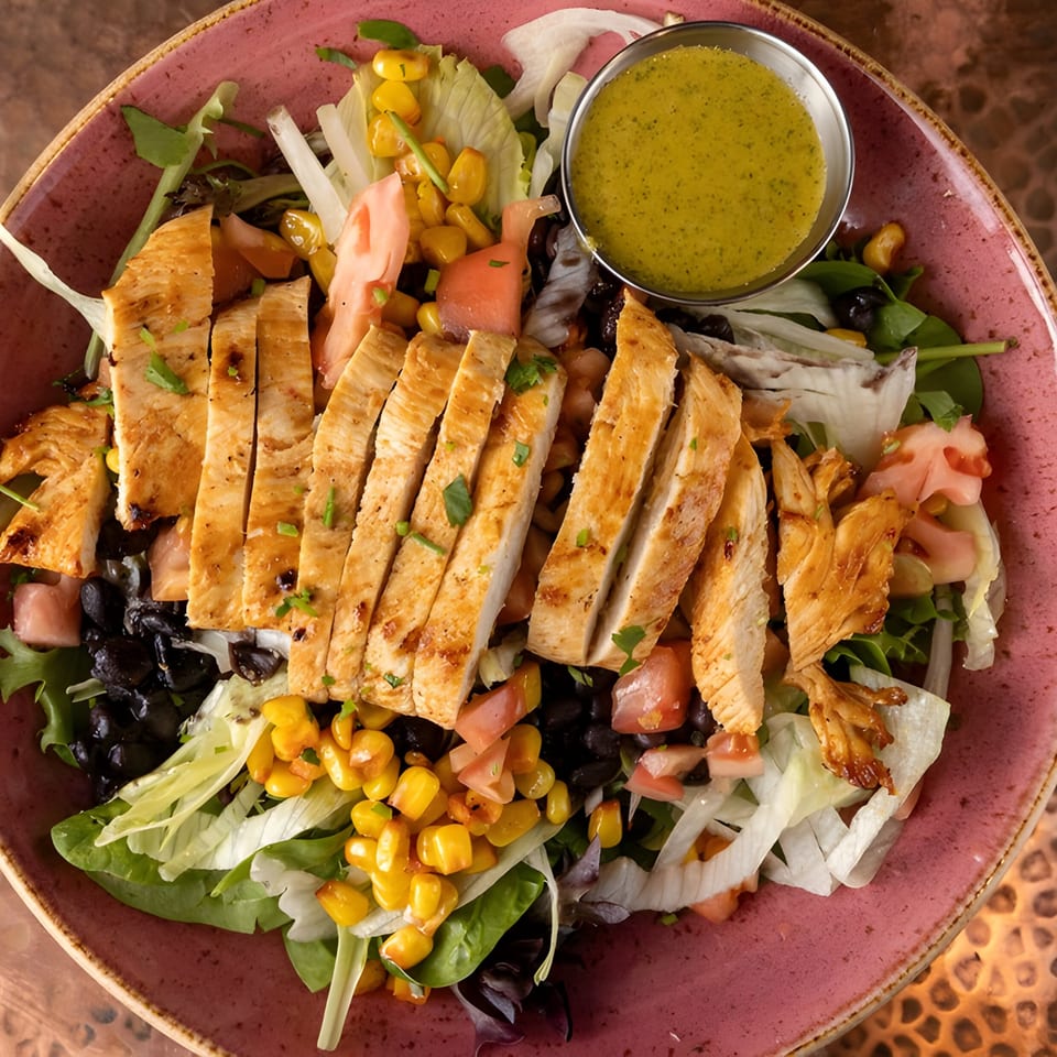 Grilled chicken salad with corn, black beans, tomatoes, and green dressing in a pink bowl.