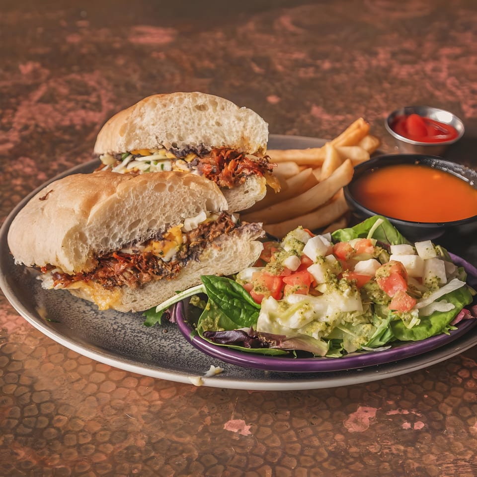Mexican torta sandwich with shredded meat, fries, a side salad, and dipping sauces on a plate.