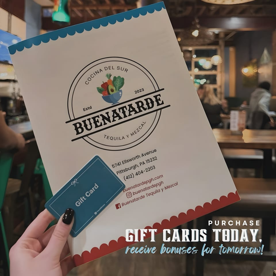 Hand holding a Buenatarde gift card and menu, with a blurred restaurant interior in the background.