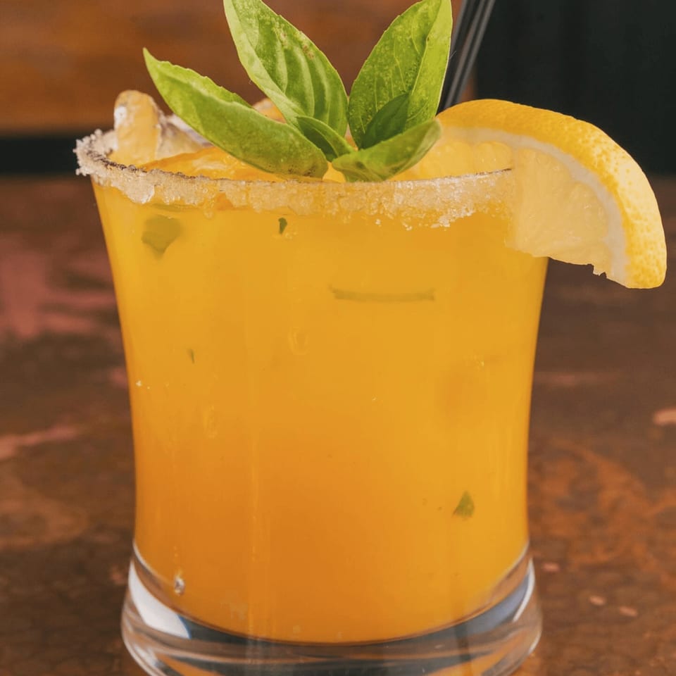 A vibrant orange cocktail with a sugar-rimmed glass, garnished with fresh basil and a lemon slice.