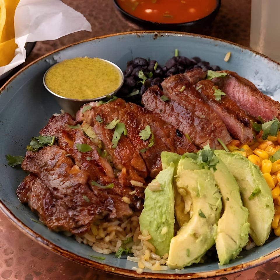 A Mexican steak bowl with rice, black beans, corn, avocado slices, and a side of green sauce.