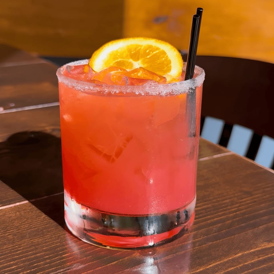 A vibrant red Buenatarde Tequila y Mezcal cocktail with an orange slice and black straws.